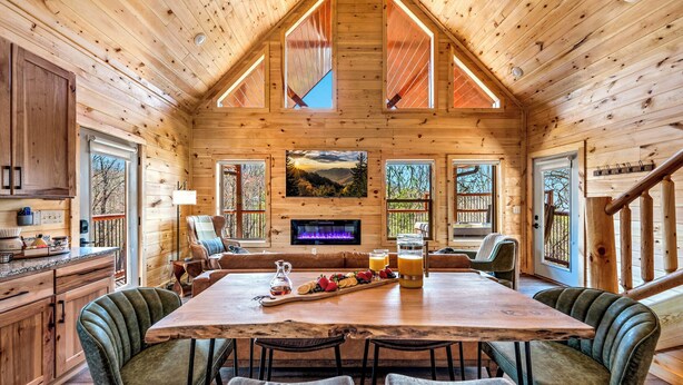 A wooden cabin interior with a large dining table, cushioned seating, a fireplace, and large windows.