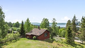 A red cabin with a tiled roof surrounded by a lush green landscape and a lake in the background. - Vacation home Alice by Interhome (Tällberg)