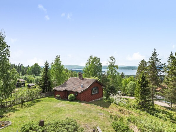 A red cabin with a tiled roof surrounded by a lush green landscape and a lake in the background.