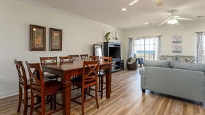 A dining area with a wooden table and chairs, a television, and a grey sofa in a living room with wooden flooring and a ceiling fan.