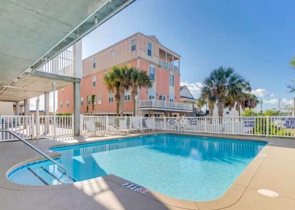 A swimming pool with a white fence, a covered area, and a building with balconies in the background.