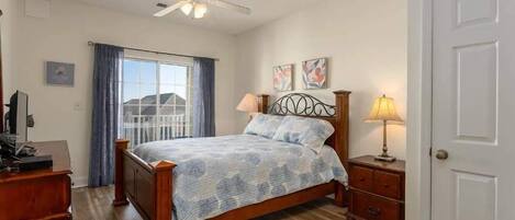 A bedroom with a bed, a desk, a chair, a ceiling fan, a window with curtains, and a door.