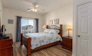 A bedroom with a bed, a desk, a chair, a ceiling fan, a window with curtains, and a door.
