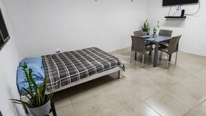 Una habitación pequeña y limpia con cama, escritorio y silla. Hay un televisor montado en la pared y una planta en maceta sobre el escritorio.