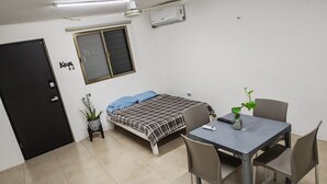 Una habitación pequeña y limpia con cama, una mesa y sillas, una planta en maceta y una puerta.