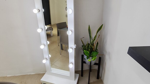 Un espejo de cuerpo entero con luces, una planta en maceta y una silla en un rincón.