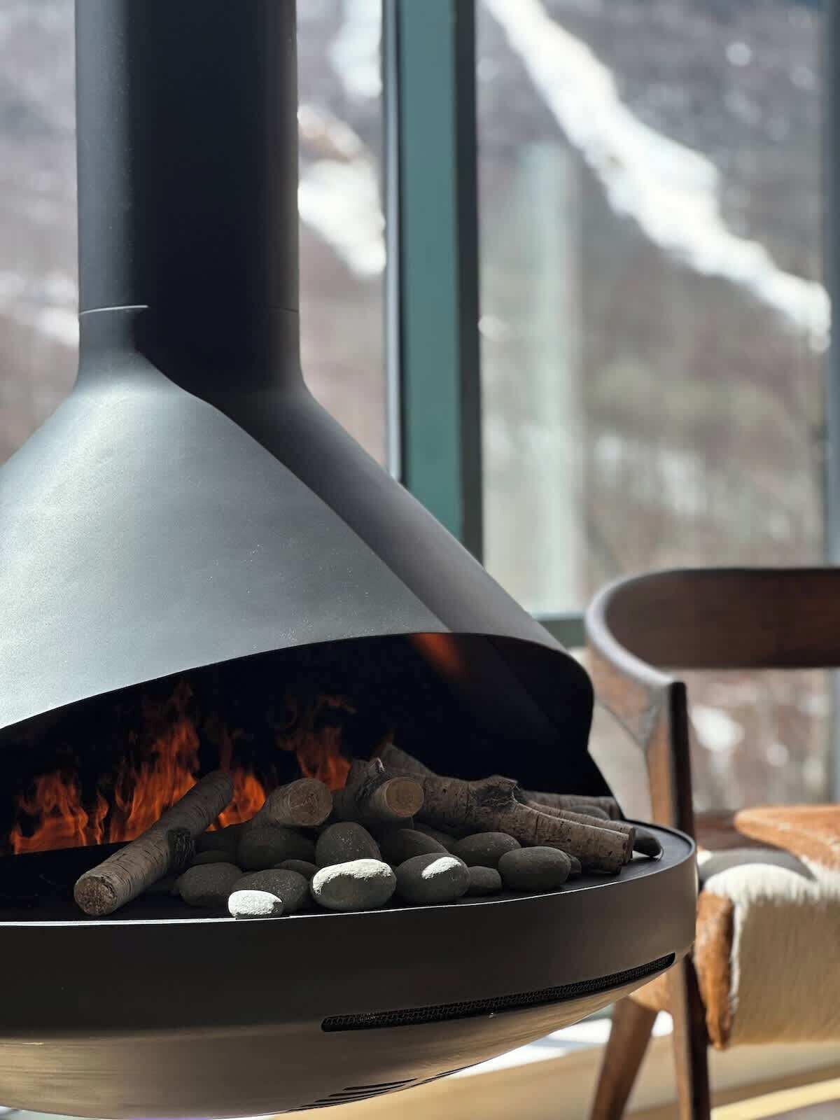 Ein moderner Kamin mit Holz und Feuer, ein Sessel und ein Fenster mit Blick auf eine verschneite Landschaft.