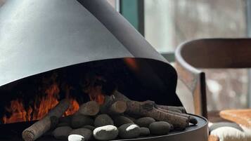 Ein moderner Kamin mit Holz und Feuer, ein Sessel und ein Fenster mit Blick auf eine verschneite Landschaft.