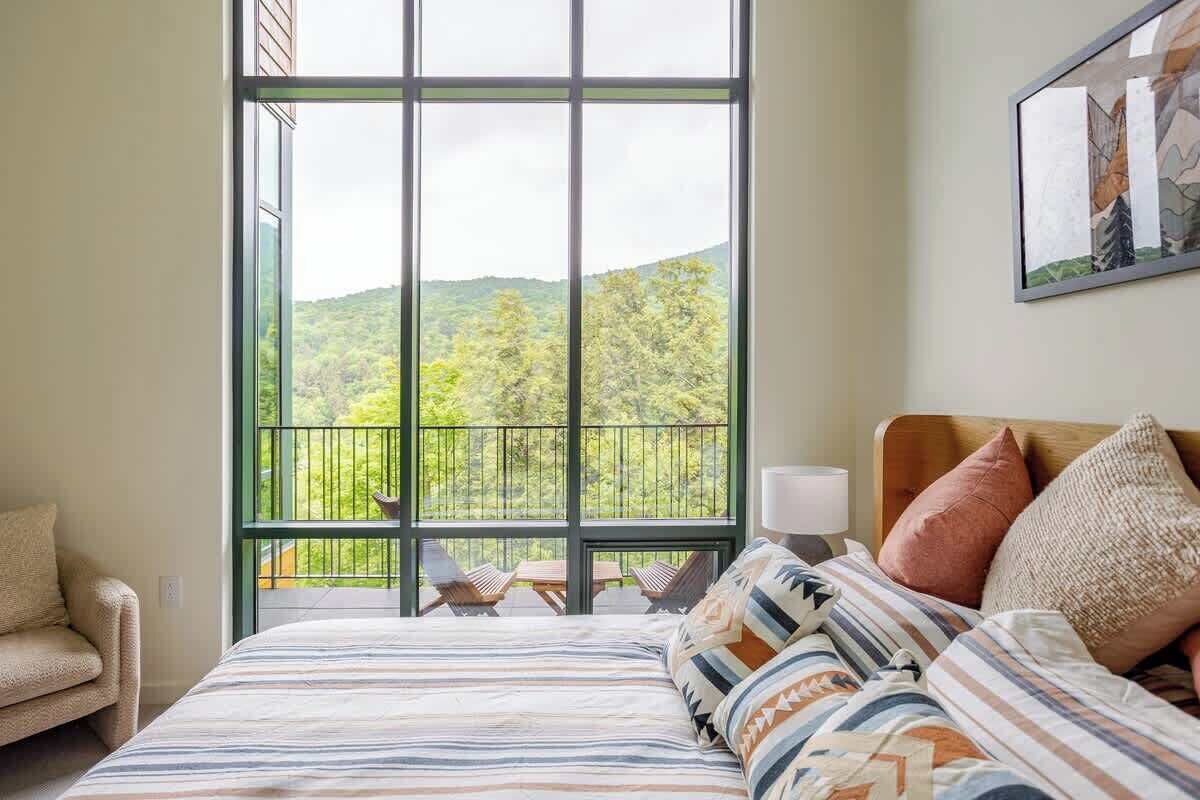A bedroom with a large window, a bed with patterned bedding, a chair, and a painting on the wall.