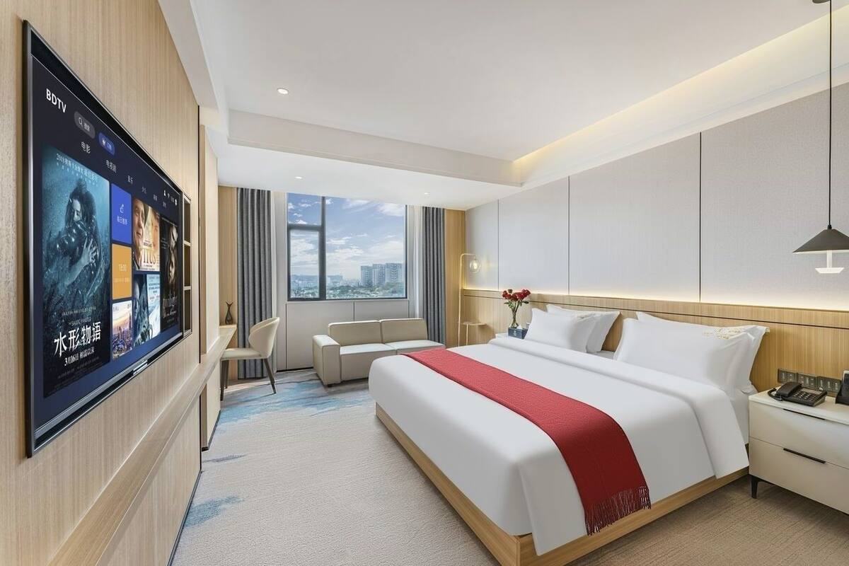 A modern hotel room with a large flat-screen TV, a bed with white bedding and a red accent, a bedside table with a phone, and a city view through the window.