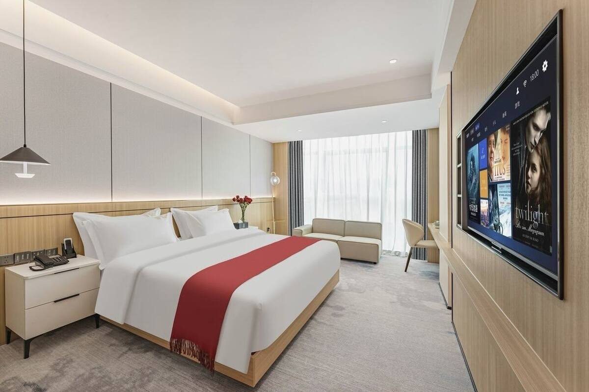 A modern hotel room with a large bed, a bedside table with a phone, a TV mounted on the wall, and a seating area with a sofa and armchairs.