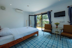 A hotel room with a bed, two chairs, a small table with a plant, a TV mounted on the wall, and a balcony with curtains. - Azure Beach Hotel (Da Nang)