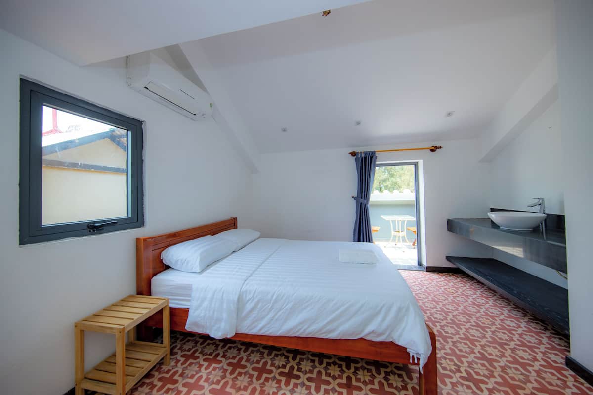 A bedroom with a bed, a small wooden stool, a window, a bathroom with a sink and a towel rack.