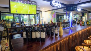 A bar area with a variety of wine bottles and glasses, a television displaying a sports game, and a neon sign above the bar. - Azure Beach Hotel (Da Nang)