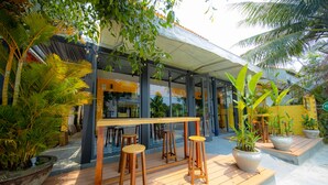 An outdoor seating area with wooden stools and potted plants, a covered patio, and a glass building in the background. - Azure Beach Hotel (Da Nang)