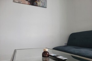 Un tavolino con piano in vetro, con un telecomando e un oggetto decorativo con la scritta 'Lounge', in una stanza con un divano scuro e un quadro appeso al muro.