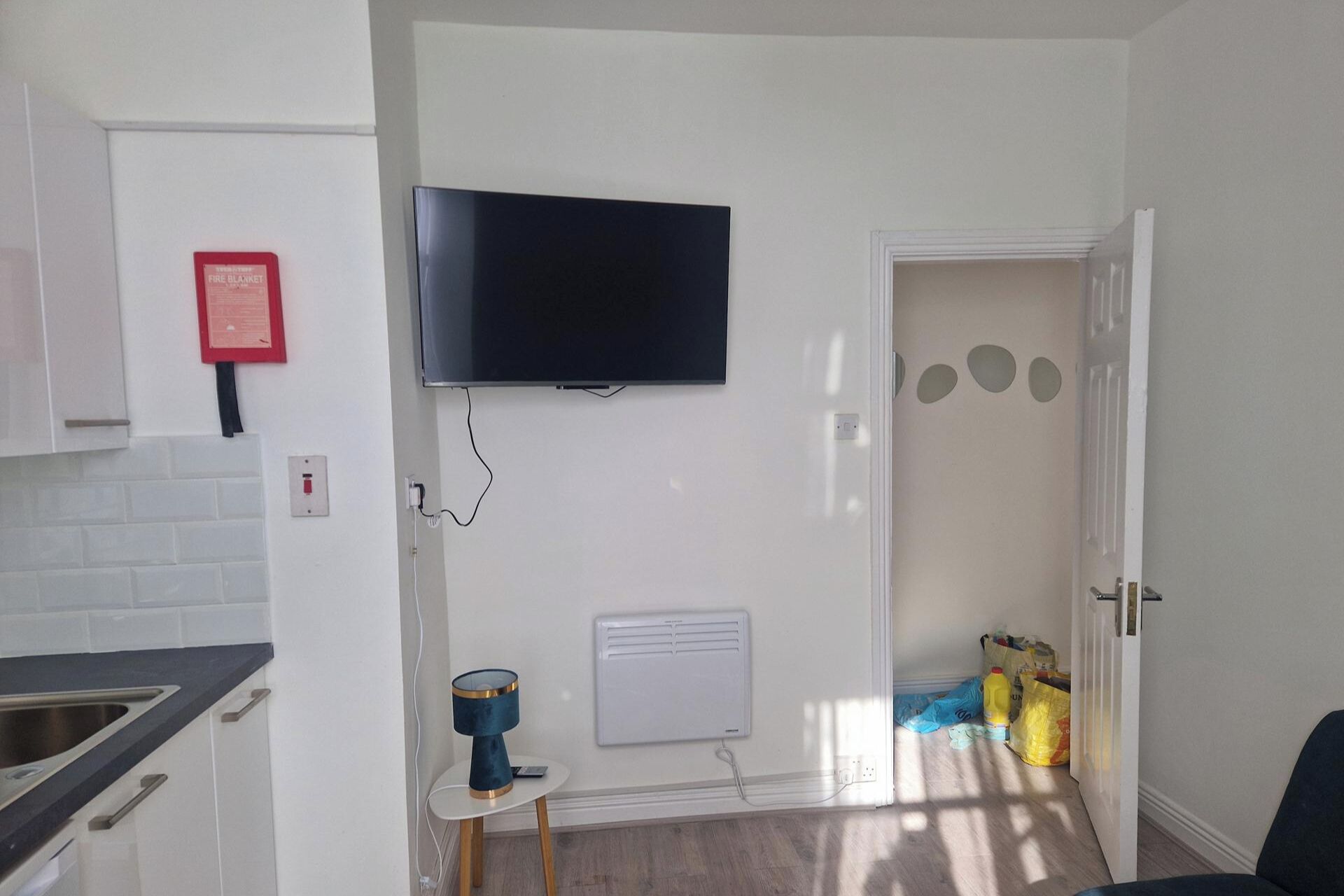 A room with a flat-screen TV mounted on the wall, a small table with a lamp, a radiator, and a door leading to another room.