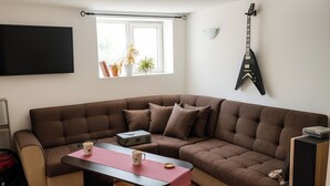 Ein modernes Wohnzimmer mit einem braunen Ecksofa, einem flachbildfernseher an der Wand, einer Gitarre und einem Fenster mit Pflanzen.