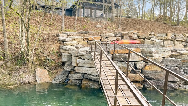 Un puente de madera cruza un cuerpo de agua transparente, conduciendo a un muro de piedra y un edificio con techo oscuro.