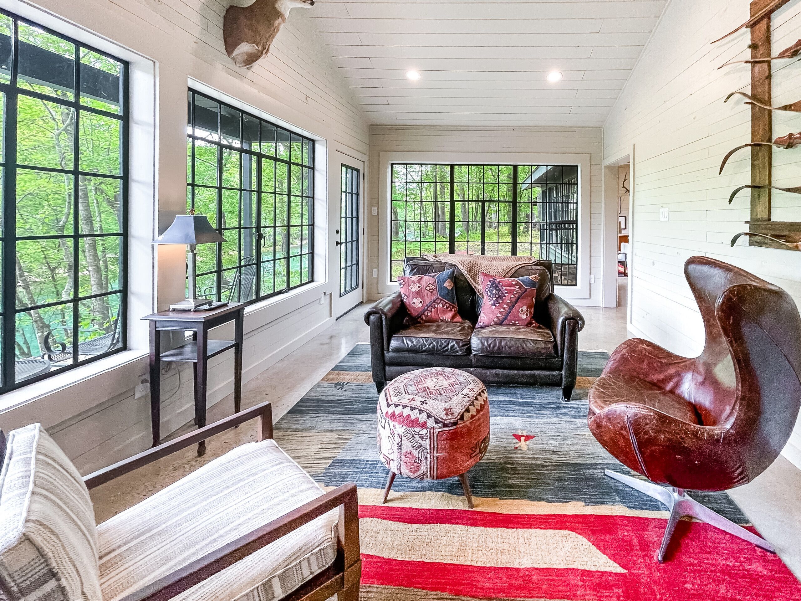 A spacious living room with a leather sofa, a red chair, and a patterned ottoman. Large windows offer a view of the greenery outside.
