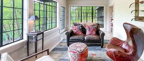 A spacious living room with a leather sofa, a red chair, and a patterned ottoman. Large windows offer a view of the greenery outside.