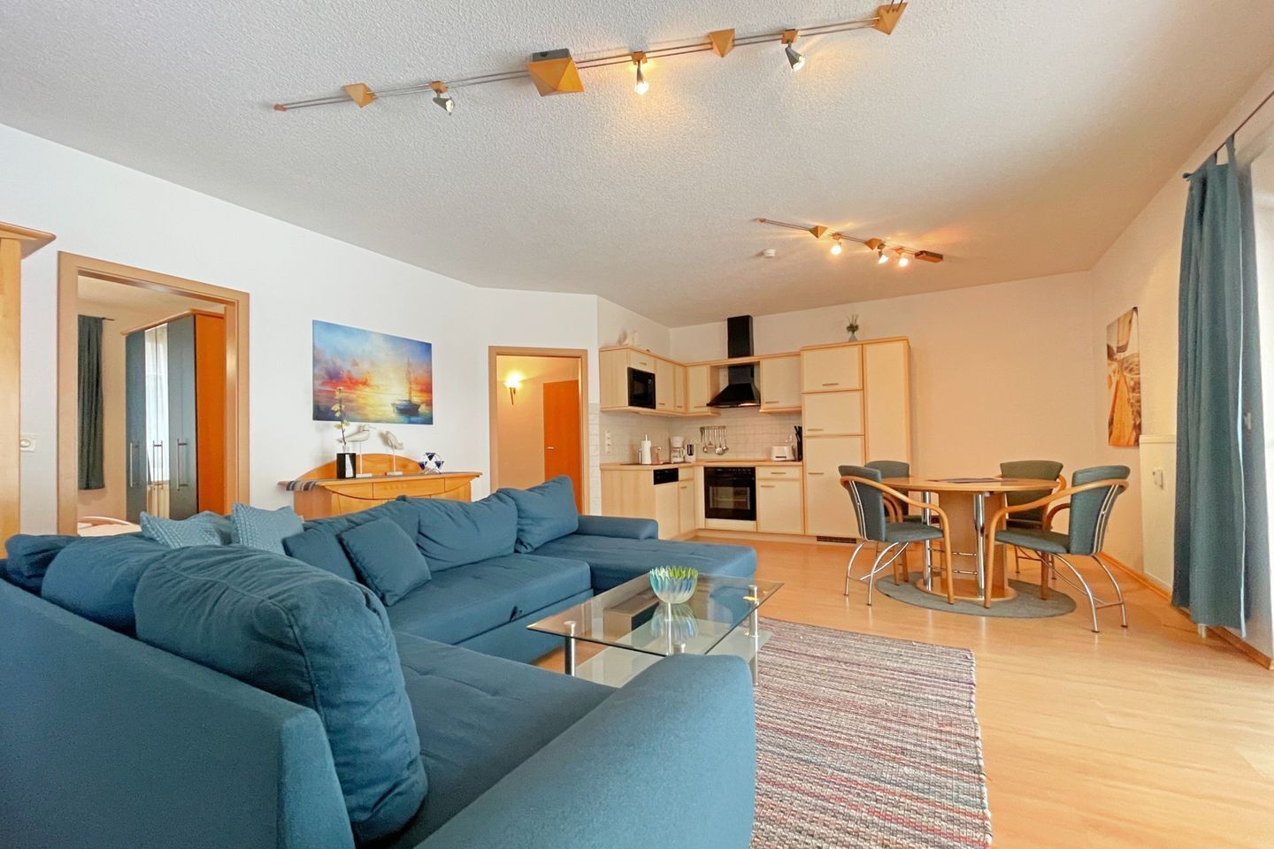 Un salon moderne avec un canapé bleu, une table basse en verre et un coin repas comprenant une table et des chaises. On aperçoit au fond une cuisine équipée de placards et d’une cuisinière.