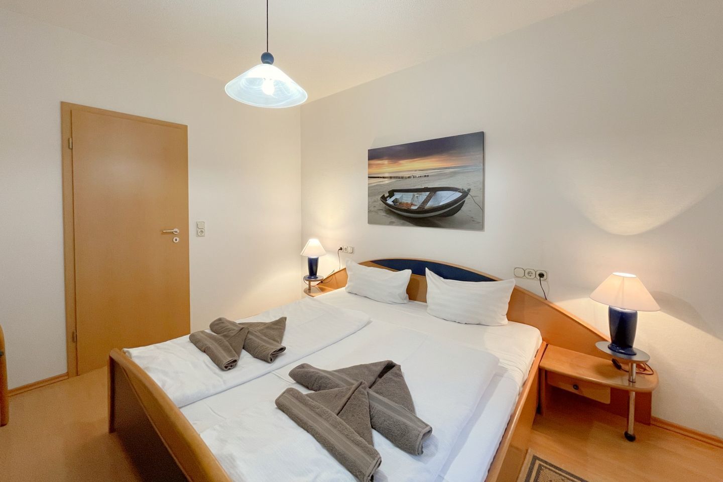 A hotel room with a double bed, two bedside tables with lamps, a painting of a boat on the wall, and a wooden door.