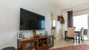 A living room with a large flat-screen TV, a wooden entertainment unit, a dining area with a table and chairs, and a view of the outside balcony. - Apartment Pardal by Interhome (Llançà)