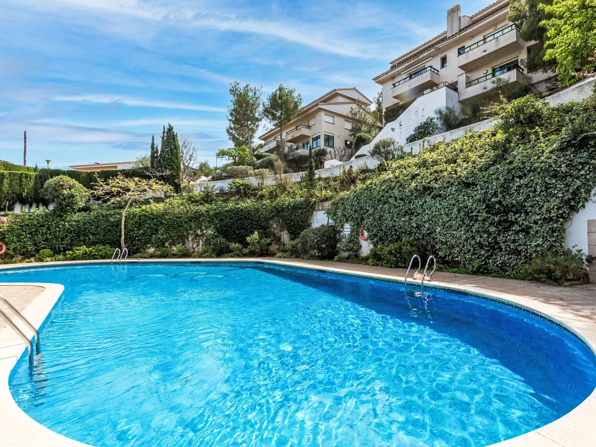 Una piscina con agua cristalina azul, rodeada de exuberante vegetación y un edificio blanco con balcones.
