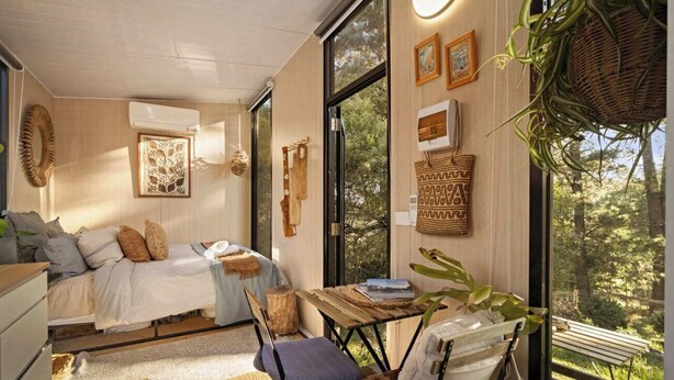 Un dormitorio acogedor con una cama, una mesita pequeña y una silla. Hay ventanas amplias, una maceta colgante y cuadros en la pared.