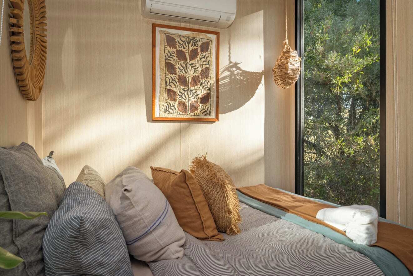 Una habitación acogedora con cama, almohadas, una obra de arte enmarcada, una lámpara colgante y vistas a árboles a través de una ventana.