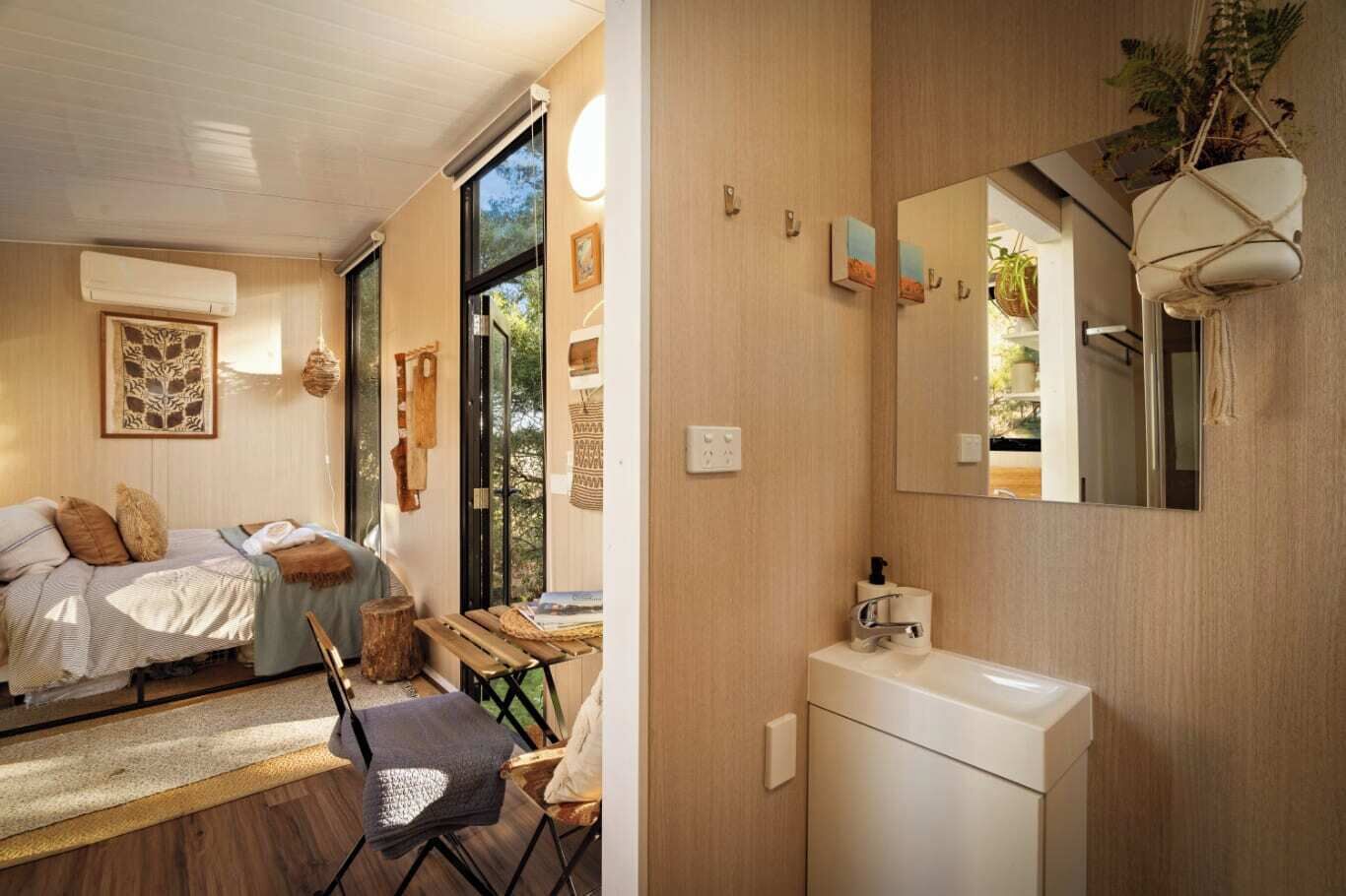 Une chambre à coucher moderne avec un lit, une chaise et un bureau. Il y a une grande fenêtre et un miroir au-dessus du lavabo dans la salle de bain.