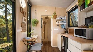 Una cocina compacta con suelo de madera, una zona de comedor con mesa y sillas, y vistas a árboles a través de amplios ventanales.