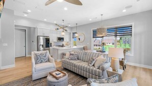 Una sala de estar moderna con sofá, sillón y reposapiés, una cocina con armarios blancos y un ventanal con persianas.