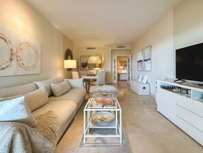A modern living room with a sofa, coffee table, TV, and artwork. - Hoyo 3 | Marbella Resort Golf Pools Apartment (Benahavís)
