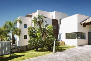 A modern two-story house with a flat roof, a stone wall, and a well-maintained lawn with palm trees. - Hoyo 3 | Marbella Resort Golf Pools Apartment (Benahavís)