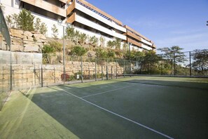 An outdoor tennis court with a stone retaining wall and a modern building with balconies in the background. - Hoyo 3 | Marbella Resort Golf Pools Apartment (Benahavís)