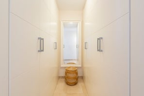 A narrow hallway with white tiled walls, a window at the end, and a woven basket on the floor.