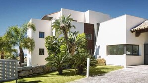 Una casa moderna de dos pisos con fachada blanca, un muro de piedra y un césped bien cuidado con palmeras.