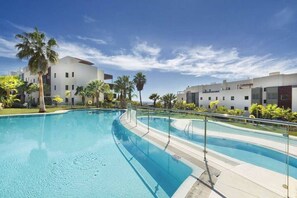A swimming pool with clear blue water, surrounded by palm trees and modern buildings. - Hoyo 3 | Marbella Resort Golf Pools Apartment (Benahavís)