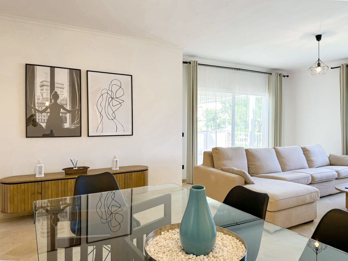 A modern living room with a glass dining table, beige sofa, and two framed artworks on the wall.