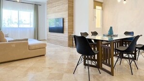 A modern living room with a beige sofa, a glass dining table with black chairs, a flat-screen TV mounted on a wooden wall, and a ceiling light fixture. - Las Brisas | Penthouse Rooftop Terrace Pool Family Only (Marbella)