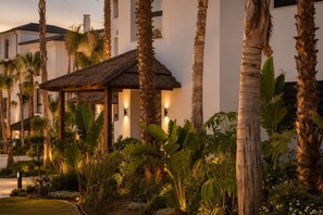 A resort with a thatched-roof structure, palm trees, and a white building with multiple windows.
