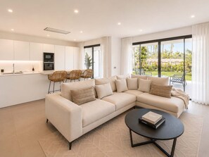 A modern living room with a sectional sofa, a dining area with chairs, a kitchen with white cabinets, and large windows with a view of greenery.