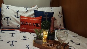 A bed with a nautical-themed bedspread, pillows with anchor designs, a small plant, a bottle of champagne, and a glass on a wooden tray. - Camper Capacidad Hasta 6 Personas (Cabo Rojo)