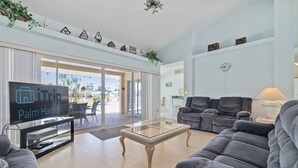 Una sala de estar moderna con un televisor de pantalla plana, sofás grises, una mesa de centro de vidrio y un amplio ventanal corredero que da a un espacio exterior.