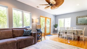 A spacious living room with a brown leather sofa, a dining area with a white table and chairs, and large windows offering a view of greenery. - Mins to Downtown in Waterfront community at Pet-Friendly Pinecrest Bungalow (Annapolis)
