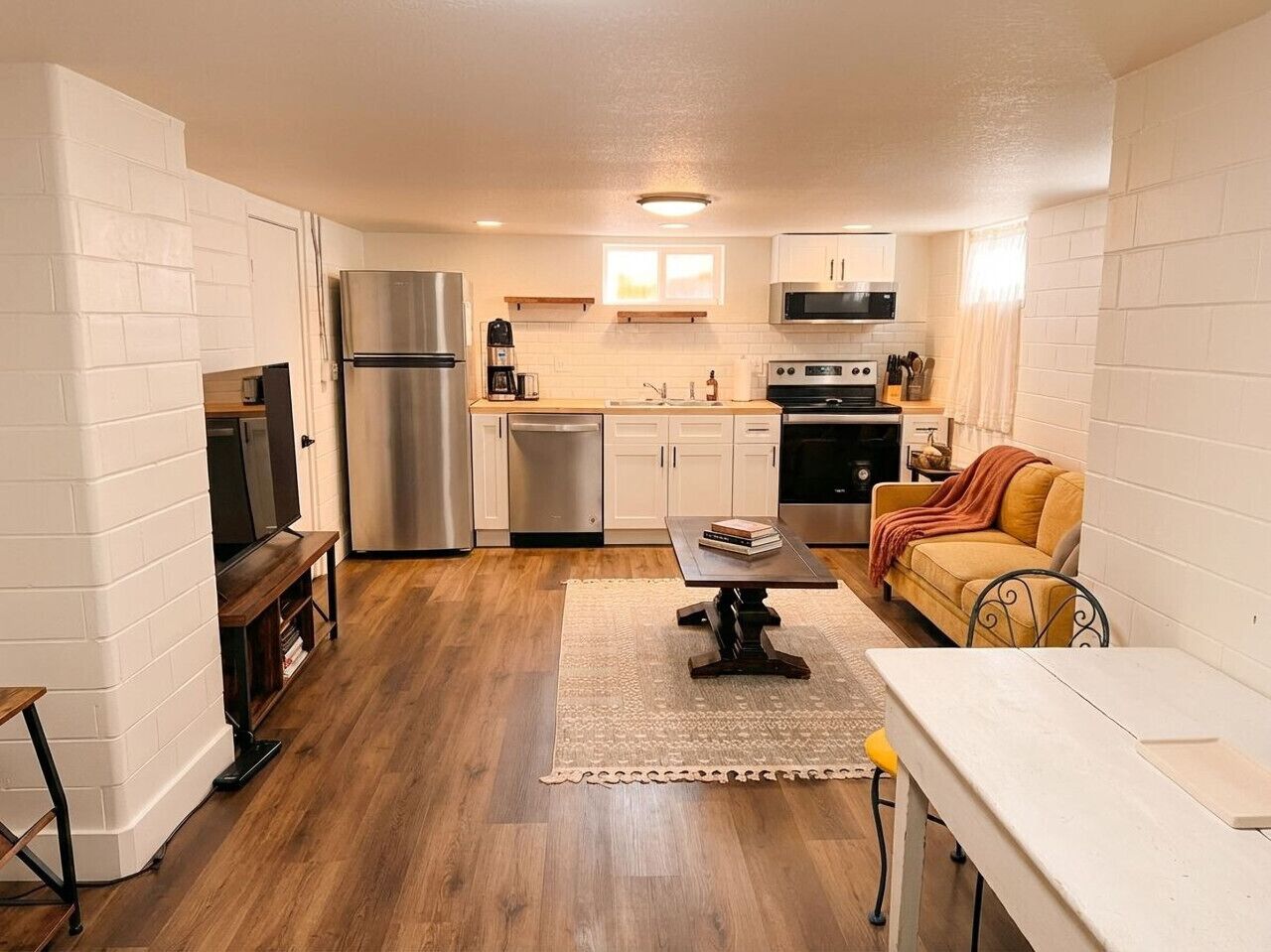 A compact kitchen with white brick walls, a wooden floor, a built-in TV, stainless steel appliances, and a yellow sofa.