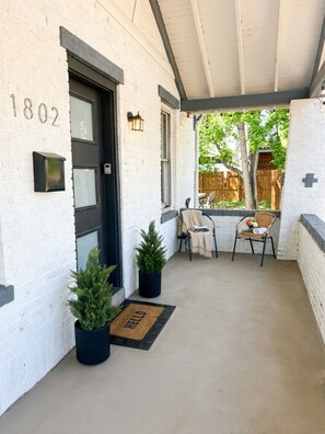 A covered porch with a door marked '1802,' two potted plants, a small table with chairs, and a 'WELCOME' mat.