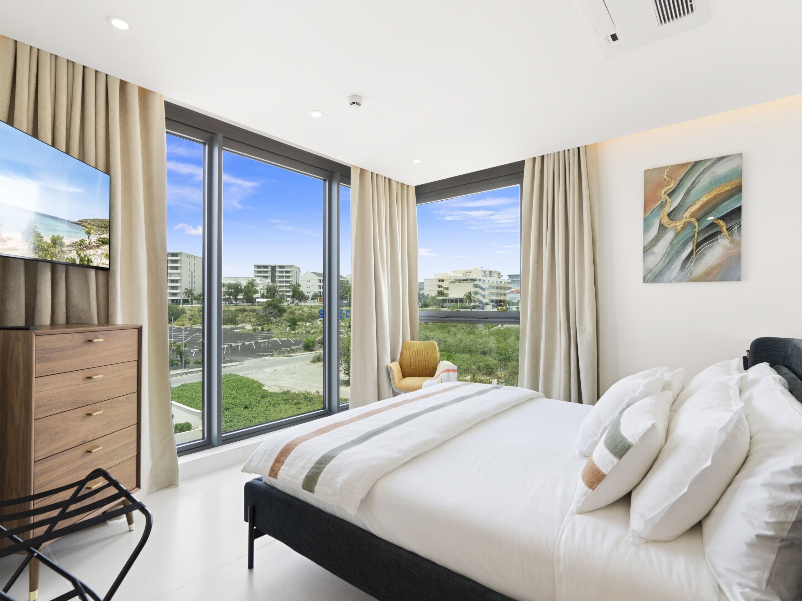 A bedroom with a large bed, a wooden dresser, a chair, and a view of a cityscape through large windows.